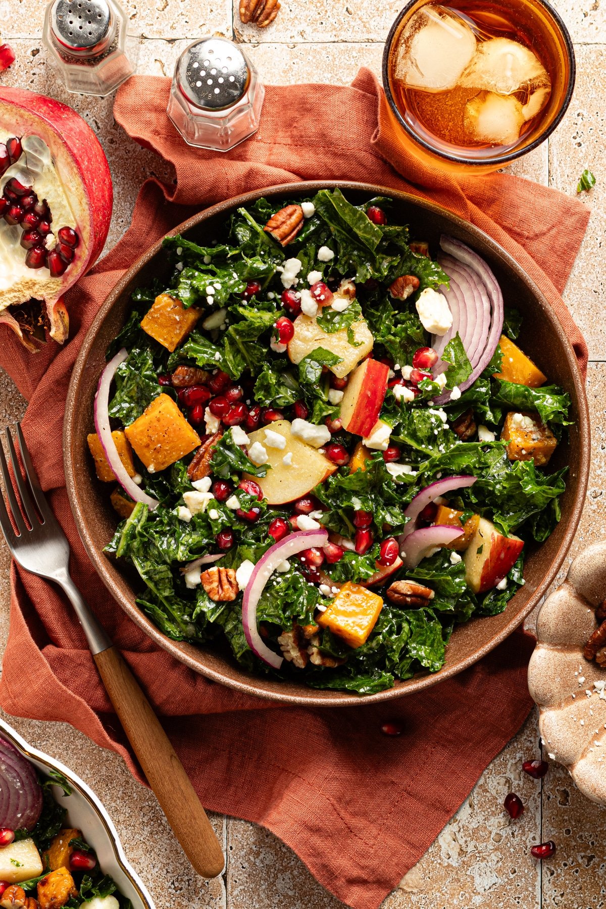 Kale butternut squash and pomegranate salad in a bowl on a table with a drink and a rust colored linen.