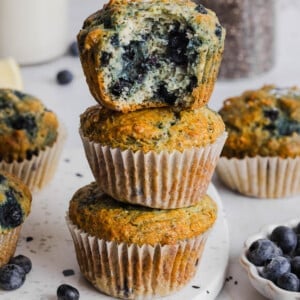 Three blueberry chia muffins sitting on top of each other in a stack on a white cutting board. The top muffin has a bite taken out of it so you can see the insides.