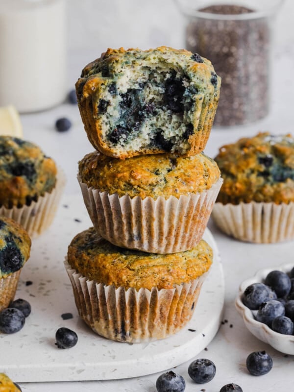 Three blueberry chia muffins sitting on top of each other in a stack on a white cutting board. The top muffin has a bite taken out of it so you can see the insides.
