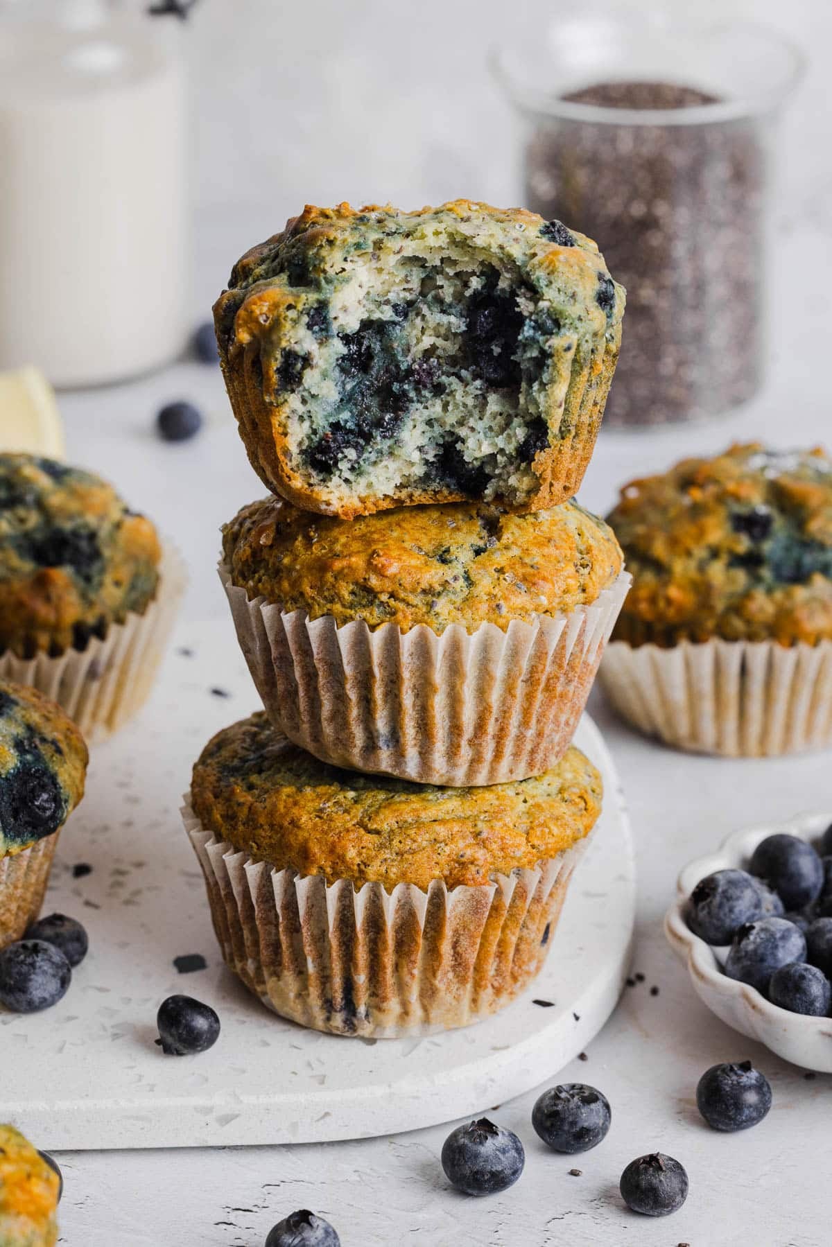 Three blueberry chia muffins sitting on top of each other in a stack on a white cutting board. The top muffin has a bite taken out of it so you can see the insides.