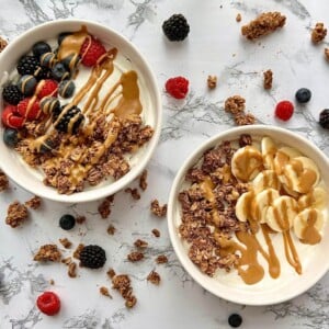 Two bowls of yogurt with granola, fruit, and drizzled peanut butter arranged on a countertop with granola and fruit scattered on the countertop.