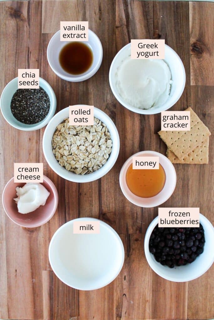 Ingredients for blueberry cheesecake overnight oats in small bowls on a wooden board with labels.