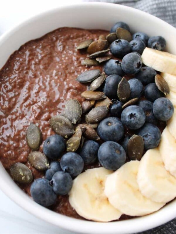 Chocolate chia seed pudding topped with banana slices, blueberries, pumpkin seeds in a white bowl on top of a countertop with a spoon and napkin.