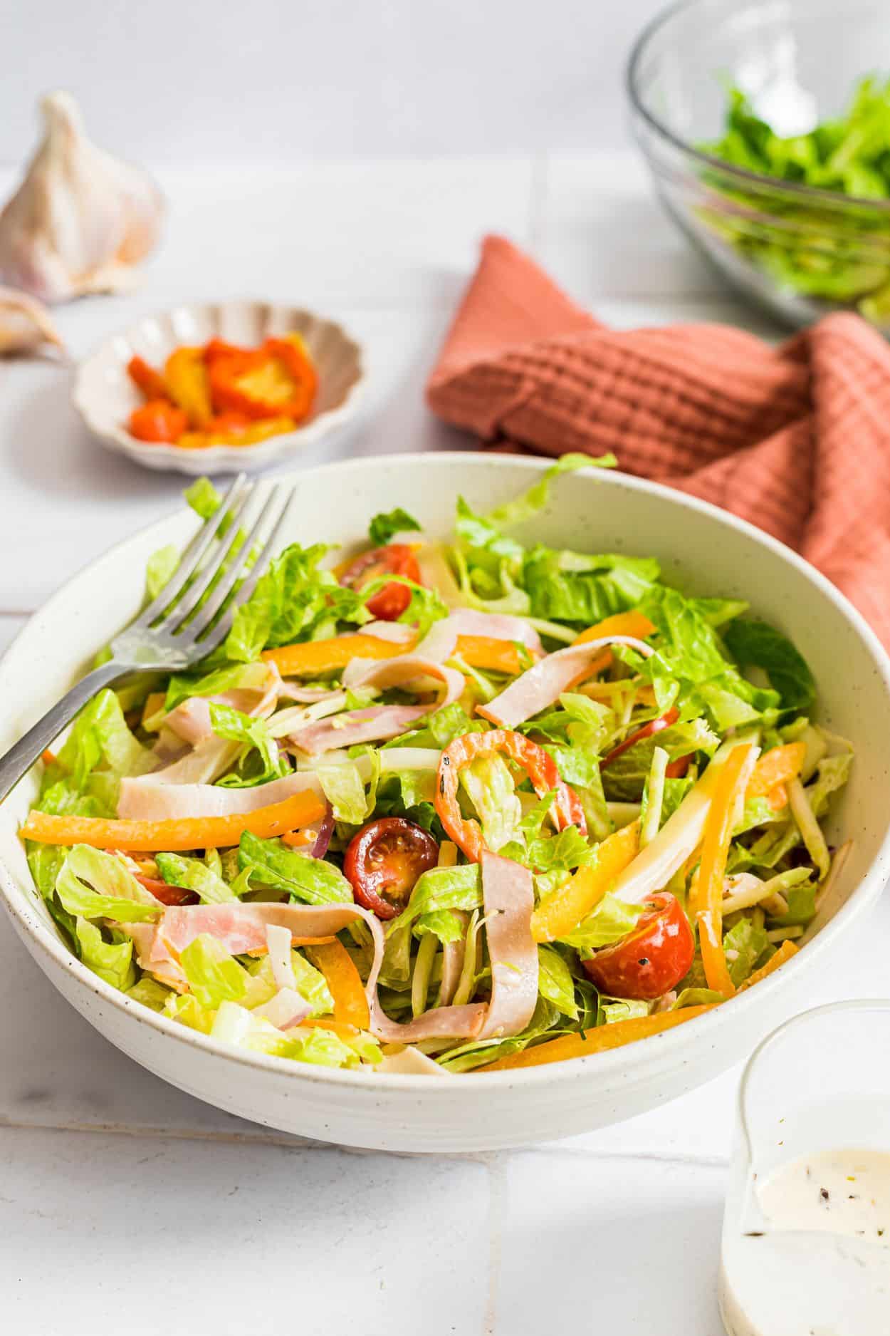 A tossed grinder salad in a white bowl on a white countertop with dressing, lettuce and banana peppers visible in the background.