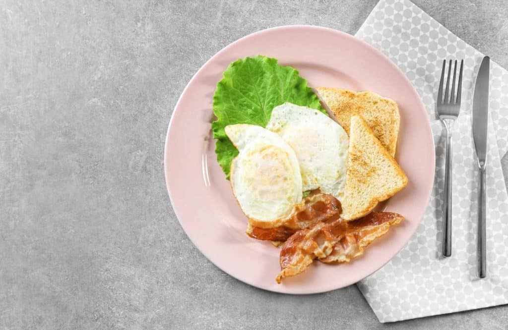 Two over hard eggs served on a plate with a slice of toast and three slices of bacon on a grey countertop and a napkin, knife, and fork.