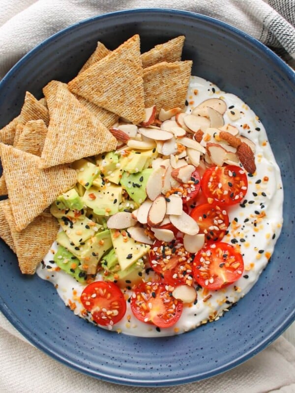 A bowl of blended cottage cheese with toppings and whole grain crackers in a blue bowl on a countertop with napkin.