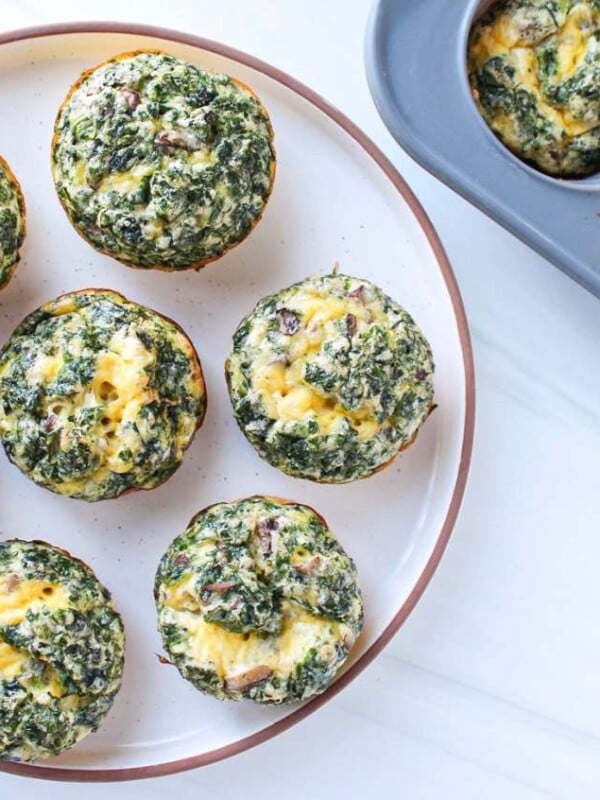 Kale and mushroom egg bites arranged on a plate with a muffin pan full of baked egg bites visible in the background.