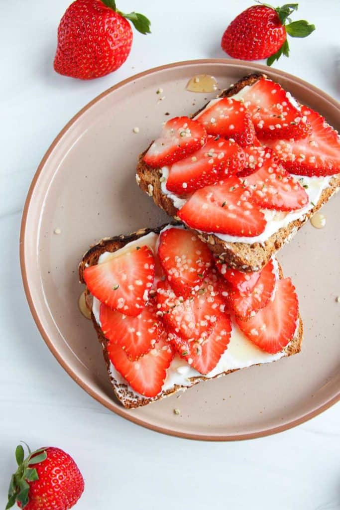 Whole grain toast with strawberries, honey and Greek yogurt plated on top of a white countertop. Strawberries are scattered on the counter.