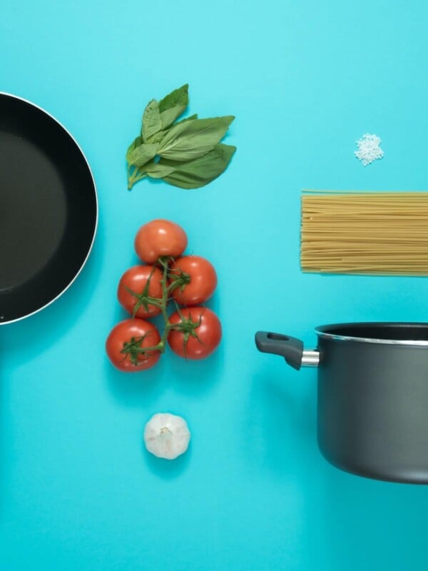 A pot and pan, fresh tomatoes, basil, and pasta on a cheerful blue background.
