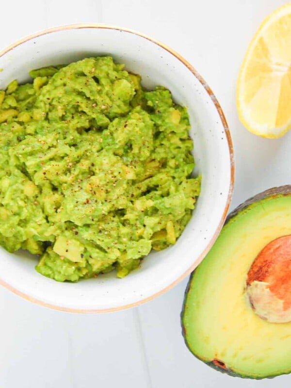A small bowl of avocado mash on a countertop, half an avocado, and a lemon wedge.