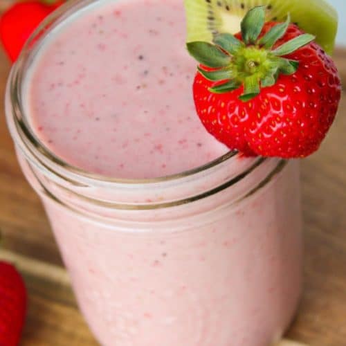 A kiwi quencher tropical smoothie in a mason jar on top of a wooden block, garnished with a strawberry and kiwi slice.