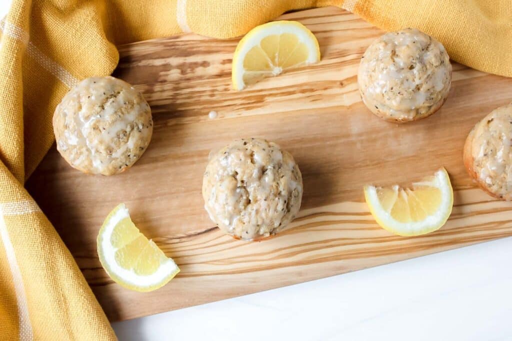 Lemon chia seed muffins arranged on a wooden serving board with lemon slices and a yellow kitchen towel.