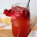 A mason jar full of iced strawberry green tea with a metal straw, strawberries and lemon visible in the background.