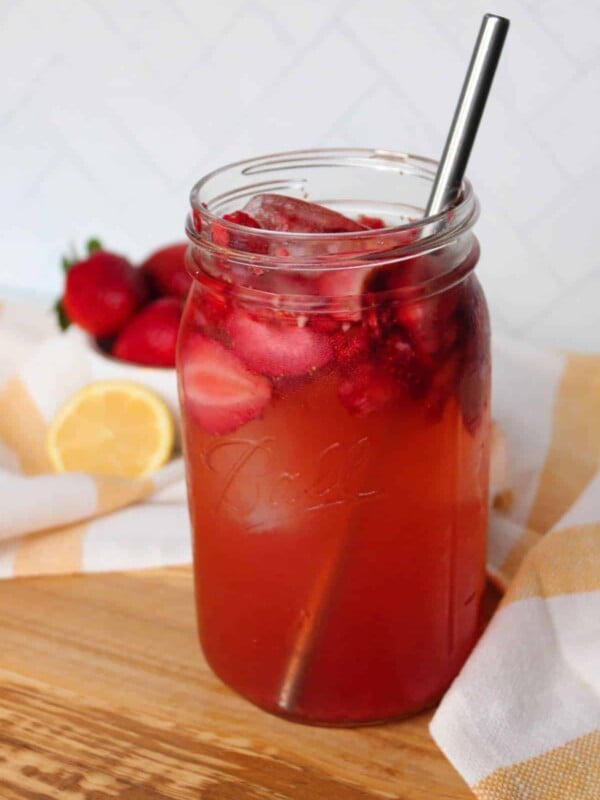 A mason jar full of iced strawberry green tea with a metal straw, strawberries and lemon visible in the background.