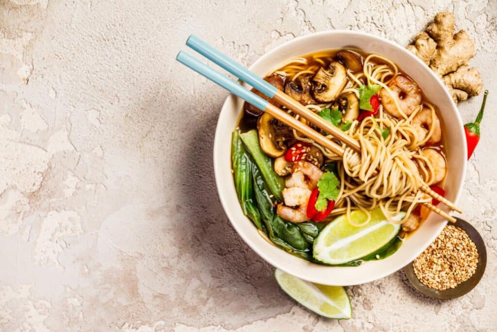 A bowl of noodles in a white bowl with chopsticks filled with mushrooms, veggies and shrimp. 