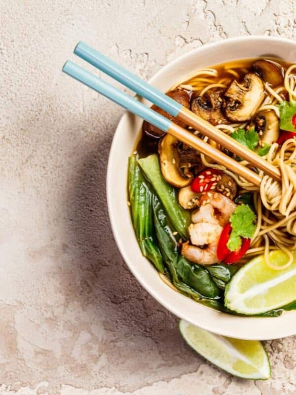 A bowl of noodles in a white bowl with chopsticks filled with mushrooms, veggies and shrimp.