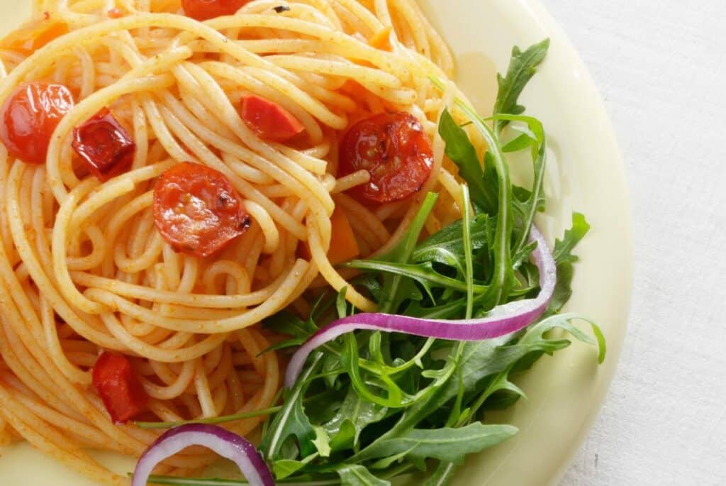 A close up shot of pasta with tomatoes and a side of greens with sliced red onion.