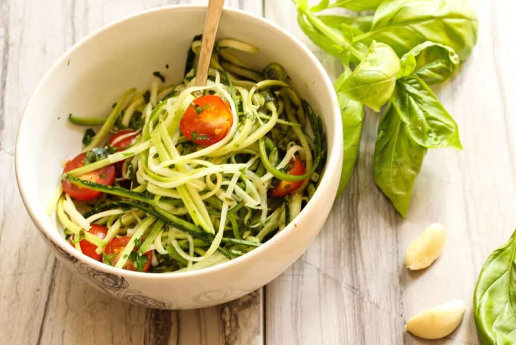 A bowl of zucchini noodles in a ceramic bowl with tomatoes on a wooden table. 