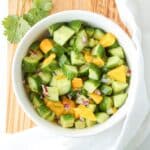 A white bowl of cucumber mango salad on a wooden board with a white kitchen towel.