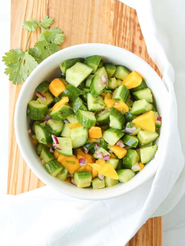 A white bowl of cucumber mango salad on a wooden board with a white kitchen towel.