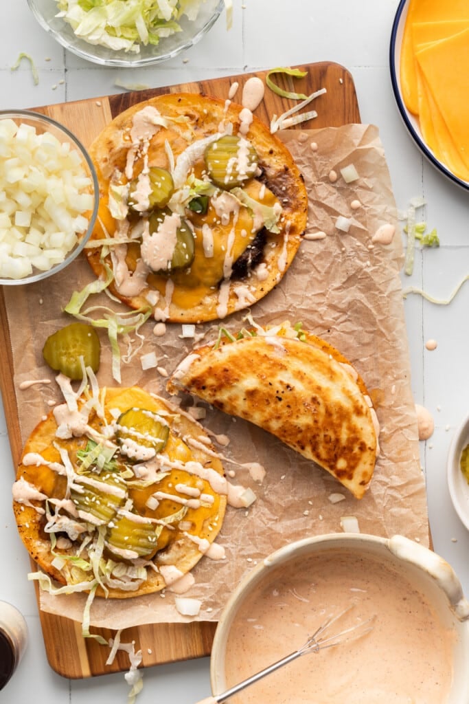 Smash burger tacos on parchment paper drizzled with dressing and topped with toppings.