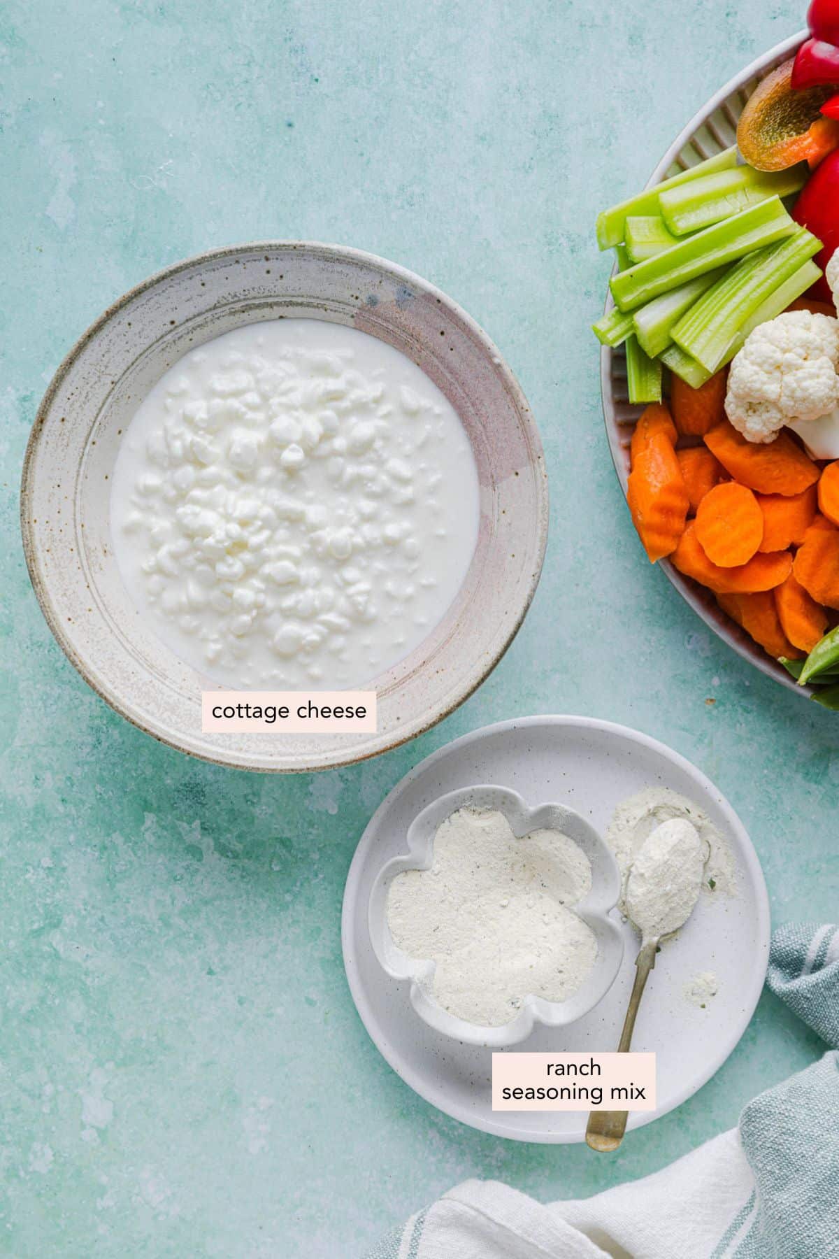 The cottage cheese and ranch seasoning mix in bowls on a light blue countertop with a plate of veggies off to the side.
