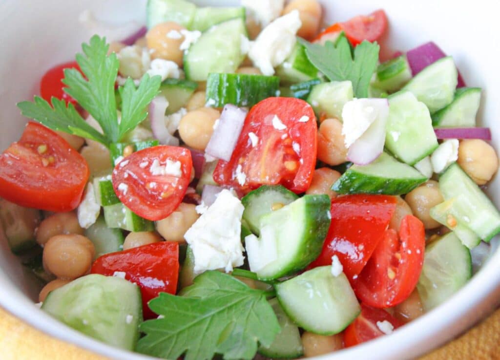 A close up shot of cucumber chickpea salad with tomatoes, red onions, and feta cheese garnished with parsley.