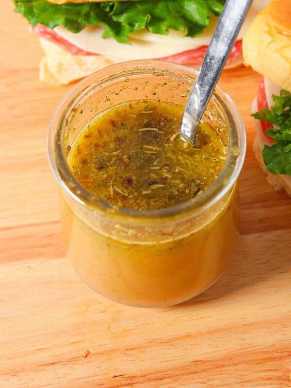 A small glass jar of italian submarine dressing with a spoon and sandwiches.