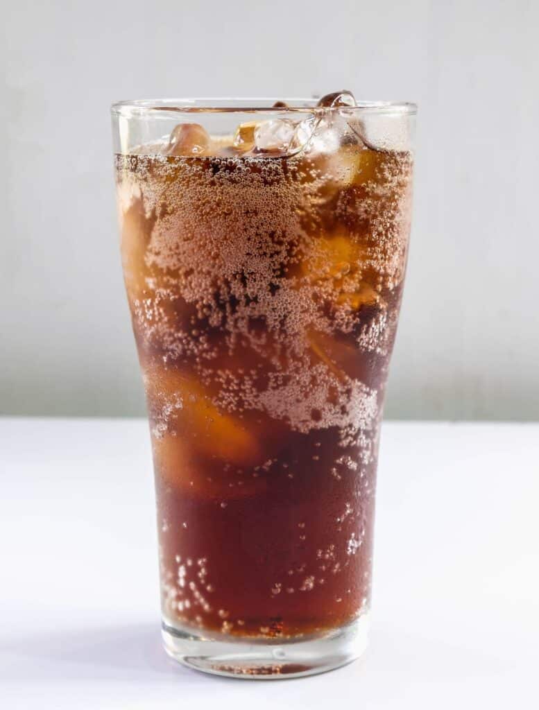 A glass full of soda with ice on a white countertop.