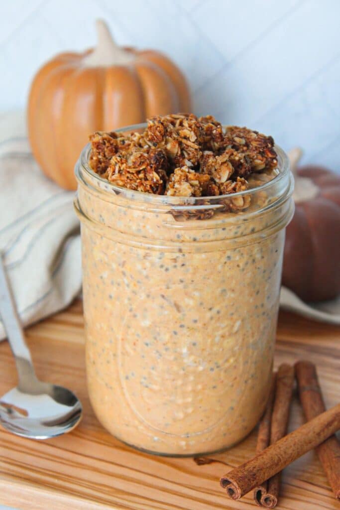 A mason jar full of pumpkin flavored overnight oats topped with granola and on a wooden board with pumpkins in the background and a spoon on the side.