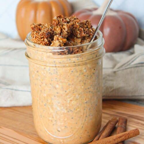 A mason jar full of pumpkin flavored overnight oats topped with granola and on a wooden board with pumpkins in the background.