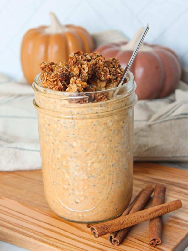 A mason jar full of pumpkin flavored overnight oats topped with granola and on a wooden board with pumpkins in the background.