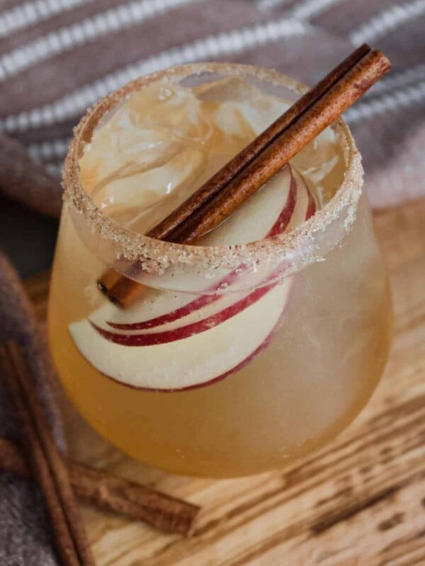 A stemless wine glass with a cinnamon sugar rim full of a sparkling apple cider mocktail garnished with apple slices and a cinnamon stick.