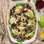 A large white serving dish filled with an arugula and pear salad with toasted pecans, feta cheese, dried cranberries and red onion.