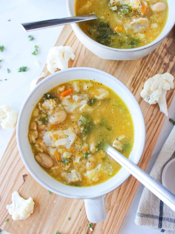 Two mugs of chicken cauliflower soup on a wooden board with cauliflower florets scattered on the countertop.