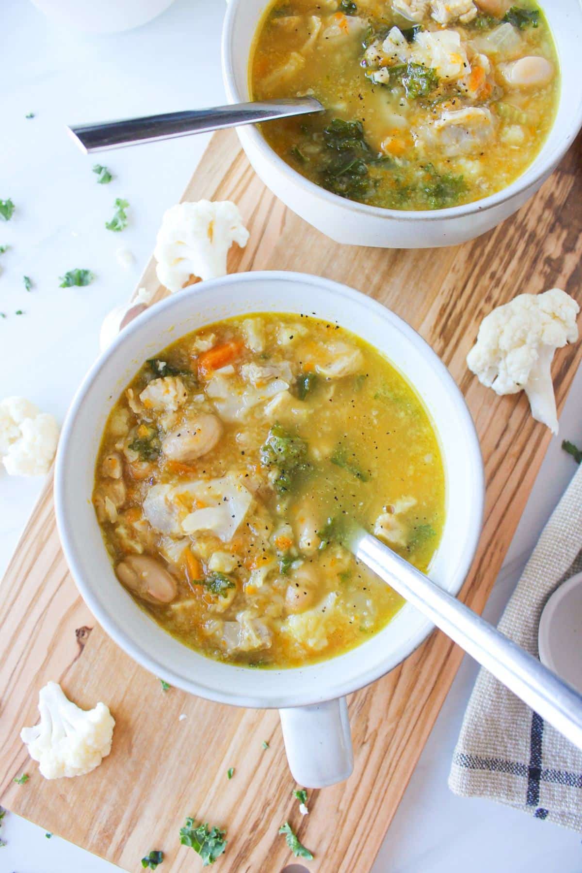 Two mugs of chicken cauliflower soup on a wooden board with cauliflower florets scattered on the countertop.