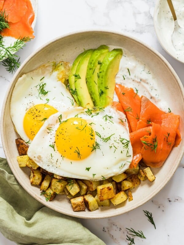 An overhead shot of a breakfast plate with salmon, sliced avocado, two fried eggs, roasted potatoes and a dill yogurt sauce.