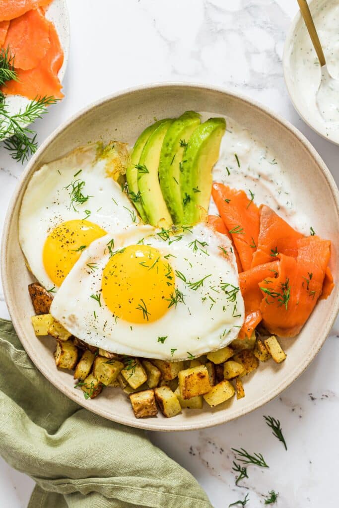 An overhead shot of a breakfast plate with salmon, sliced avocado, two fried eggs, roasted potatoes and a dill yogurt sauce.