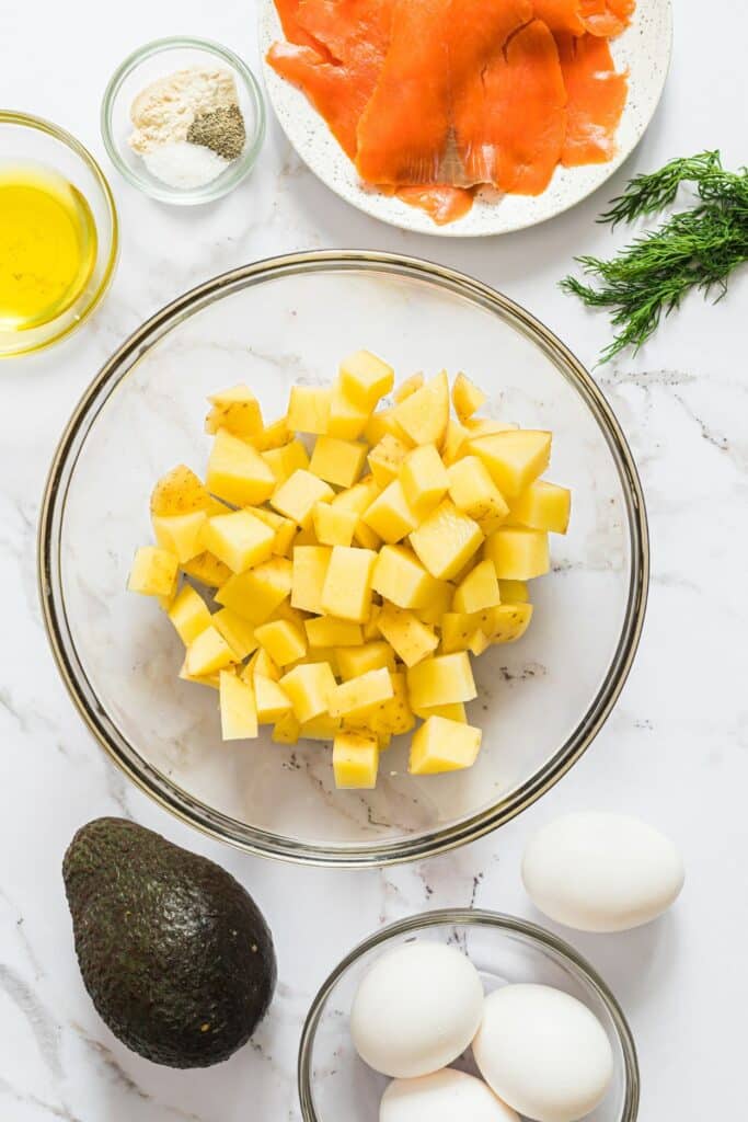 Ingredients for salmon breakfast bowls: salmon, potatoes, avocado, eggs, and ingredients for a dill yogurt sauce laid out on a white countertop and labeled.