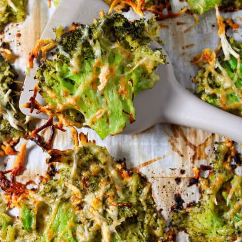 A close up shot of smashed broccoli parmesan on a sheet pan with a spatula lifting up a broccoli piece.