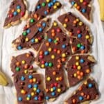 Chocolate banana bark sprinkled with mini M&Ms broken into pieces on parchment paper.