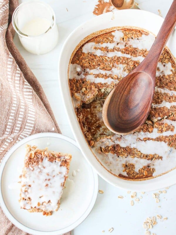 A white casserole dish with cinnamon roll oatmeal bake drizzled with a cinnamon roll glaze with a slice next to it on a plate.