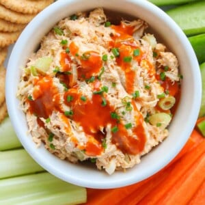 A white bowl of healthy buffalo chicken salad drizzled with buffalo sauce and garnished with chives, surrounded by veggies and crackers on a plate.