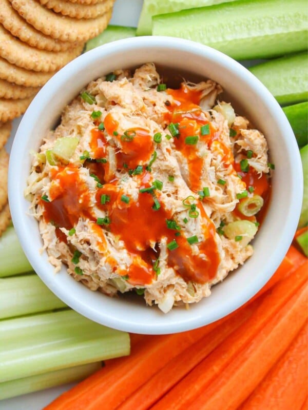 A white bowl of healthy buffalo chicken salad drizzled with buffalo sauce and garnished with chives, surrounded by veggies and crackers on a plate.