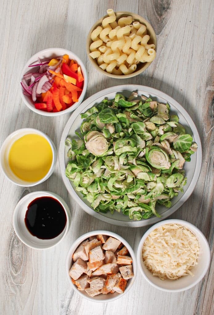 Ingredients for brussel sprout pasta salad in small bowls on a countertop: brussels sprouts, red onion, pepper, olive oil, cooked chicken, parmesan cheese, balsamic glaze, and pasta.