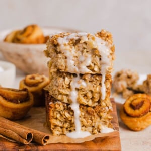 A stack of three pieces of cinnamon roll baked oats sits on a wooden cutting board with the glaze drizzle coming down the sides.