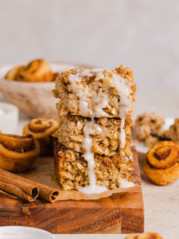 A stack of three pieces of cinnamon roll baked oats sits on a wooden cutting board with the glaze drizzle coming down the sides.