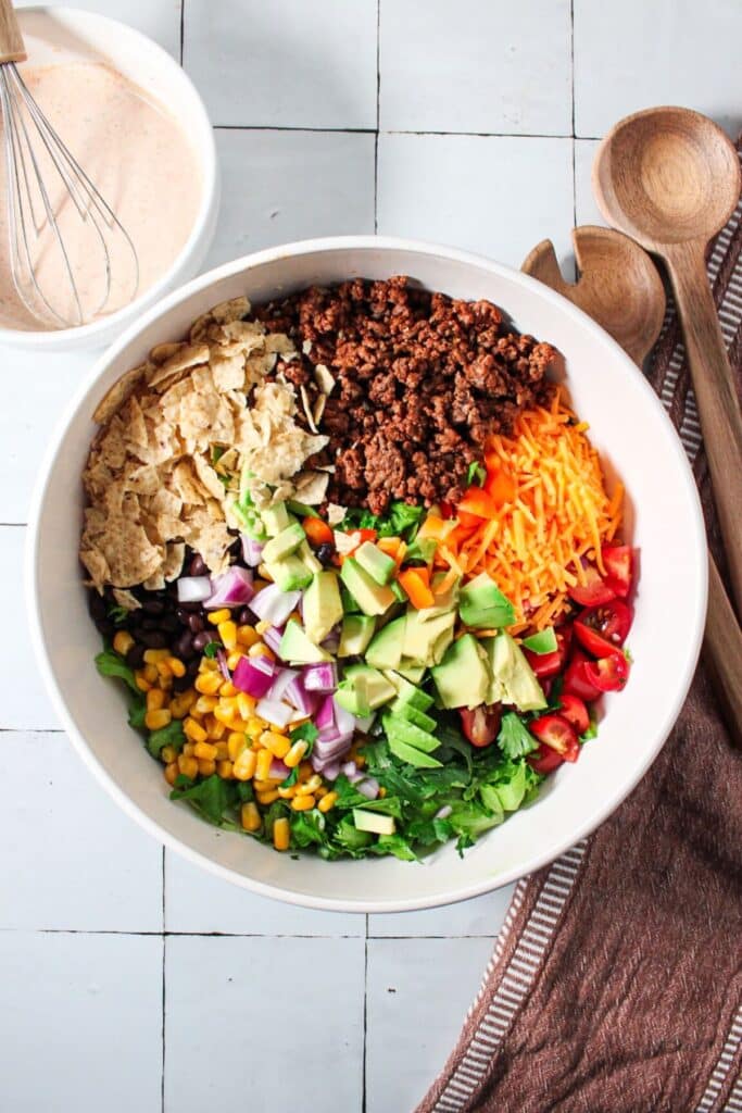Lettuce, cilantro, avocado, red onion, cheese, tomatoes, ground beef, corn, beans and chips unmixed in a bowl.