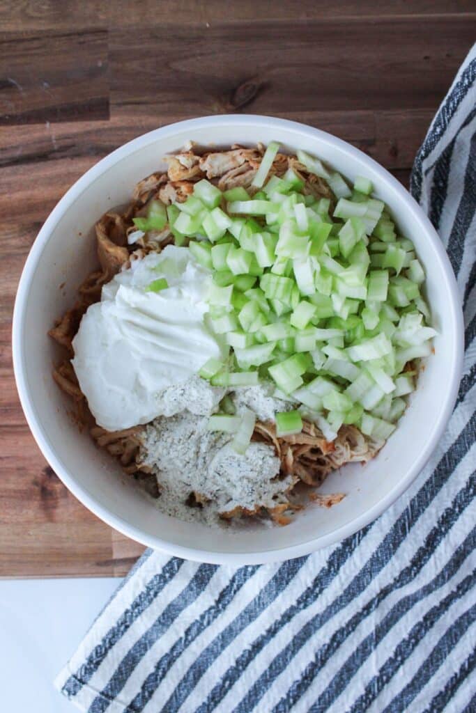 Shredded chicken, plain Greek yogurt, celery and ranch seasoning powder in a white bowl.