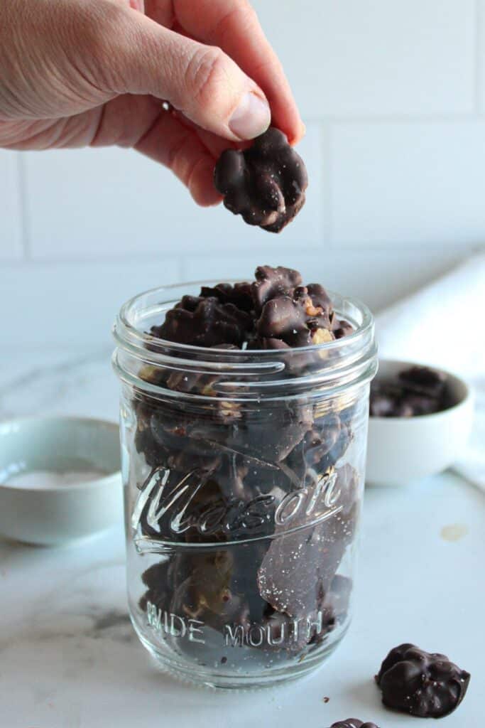 A glass jar of dark chocolate covered walnuts with a hand taking one out of the jar.